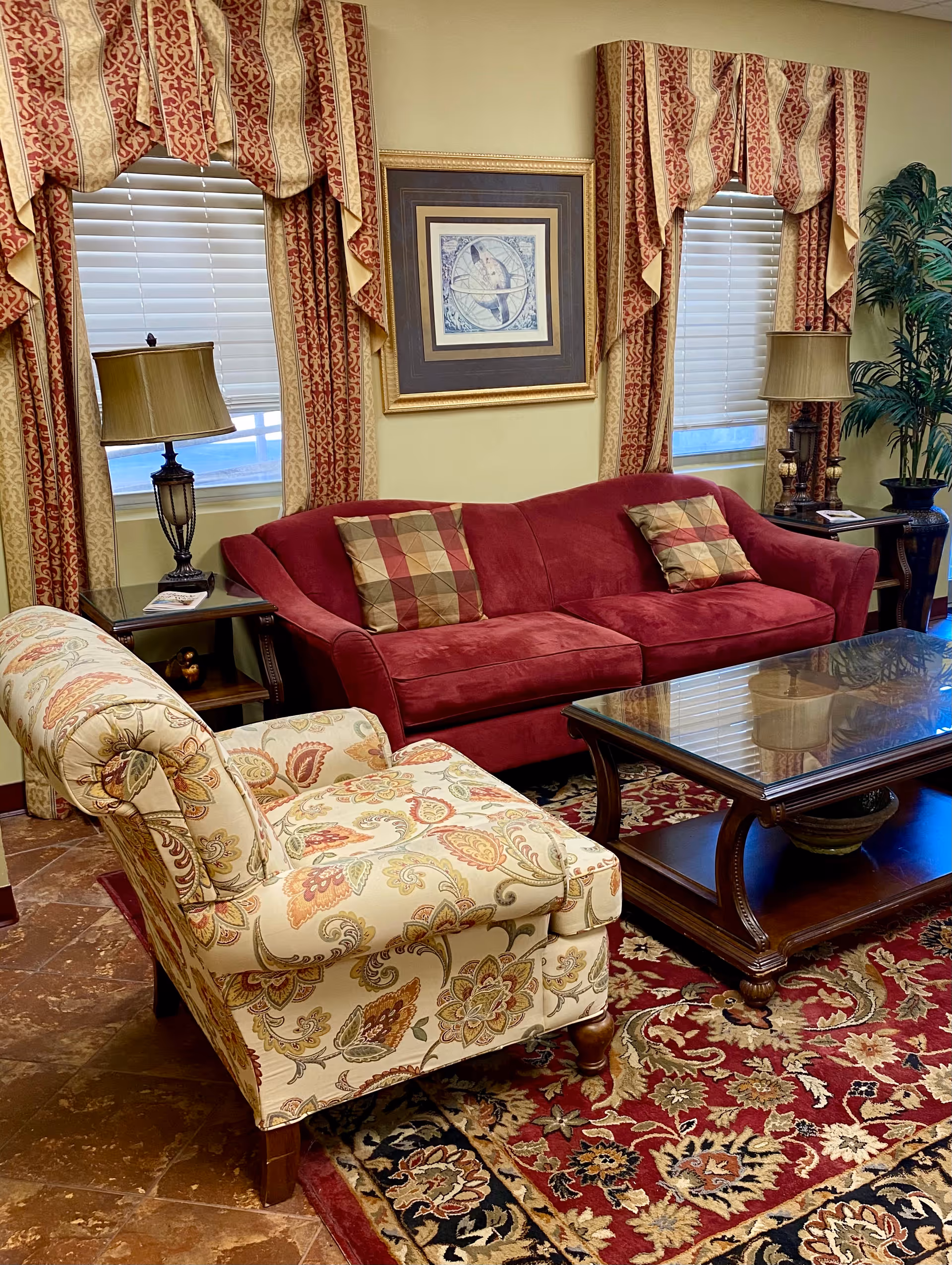 A cozy living room with a red sofa adorned with two patterned pillows, a floral upholstered armchair, a wooden coffee table with a glass top, two side tables each with a lamp, two windows with red and gold patterned curtains, a framed artwork on the wall, and a large potted plant in the corner.