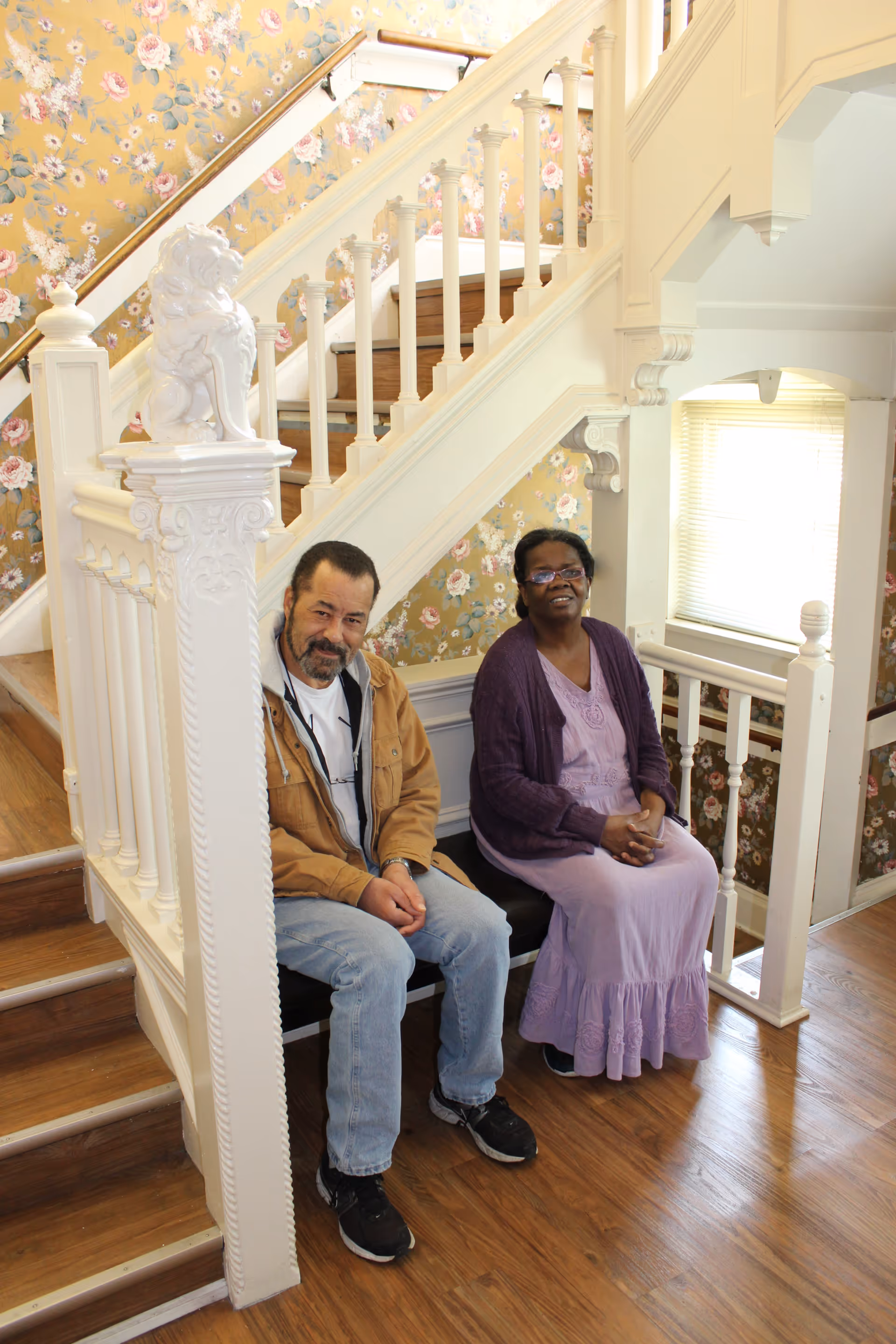 Two people sitting on a bench under a white wooden staircase with floral wallpaper in the background. The man on the left is wearing a brown jacket, white shirt, and jeans, while the woman on the right is wearing a purple dress and cardigan. The floor is wooden, and there is a window with blinds behind them.