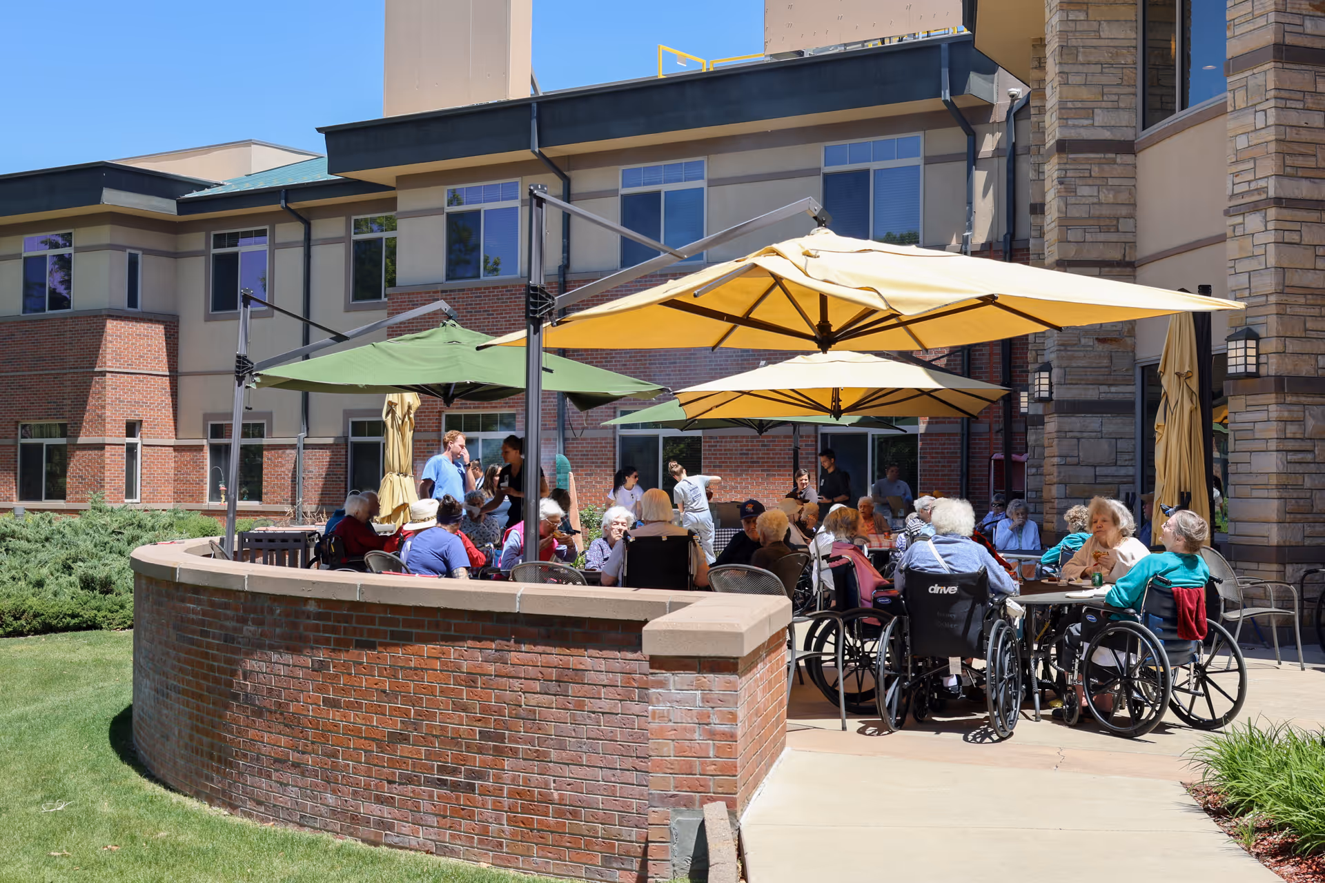 A group of elderly people, some in wheelchairs, sitting and socializing under large yellow and green patio umbrellas outside a brick and stone building on a sunny day.