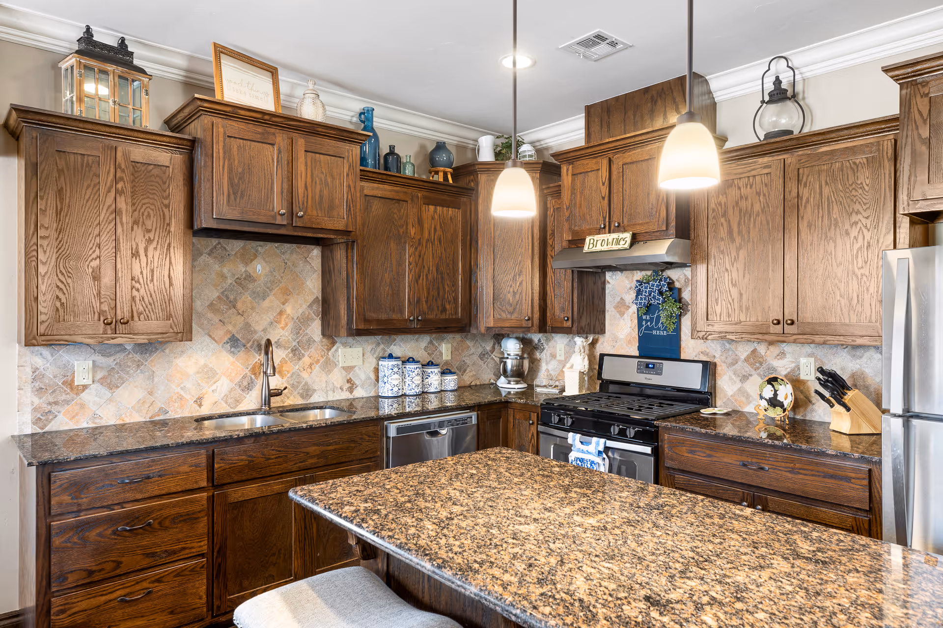 Kitchen with dark wood cabinets, granite island, pendant lights, stainless steel appliances, and a tiled backsplash.
