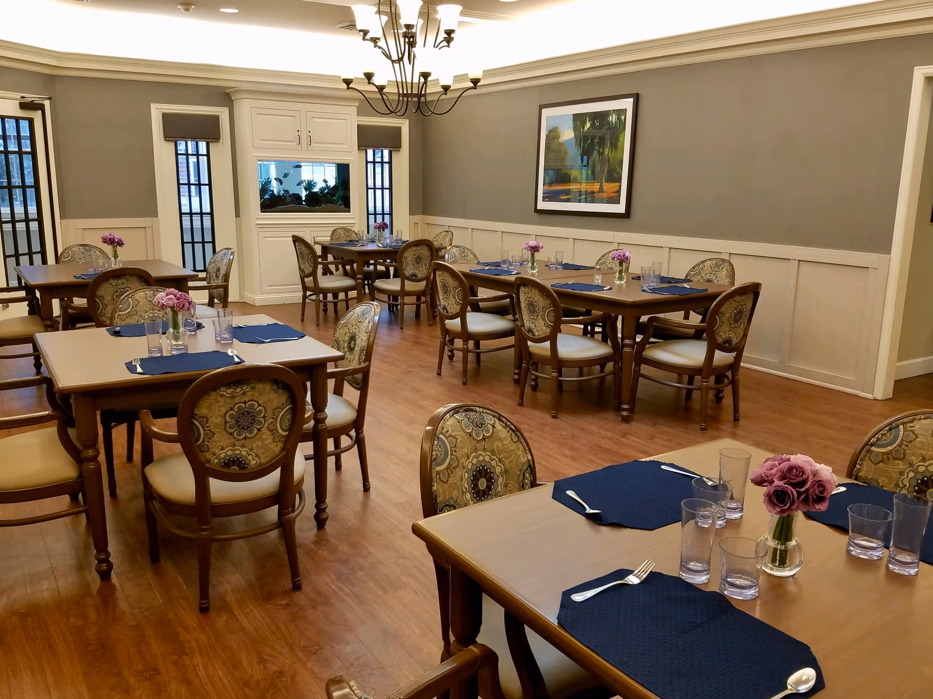 A dining room with several wooden tables and upholstered chairs arranged neatly. Each table is set with blue placemats, silverware, clear drinking glasses, and small vases with purple flowers. The room has wooden flooring, gray walls with white wainscoting, a chandelier hanging from the ceiling, and a framed painting on the wall.