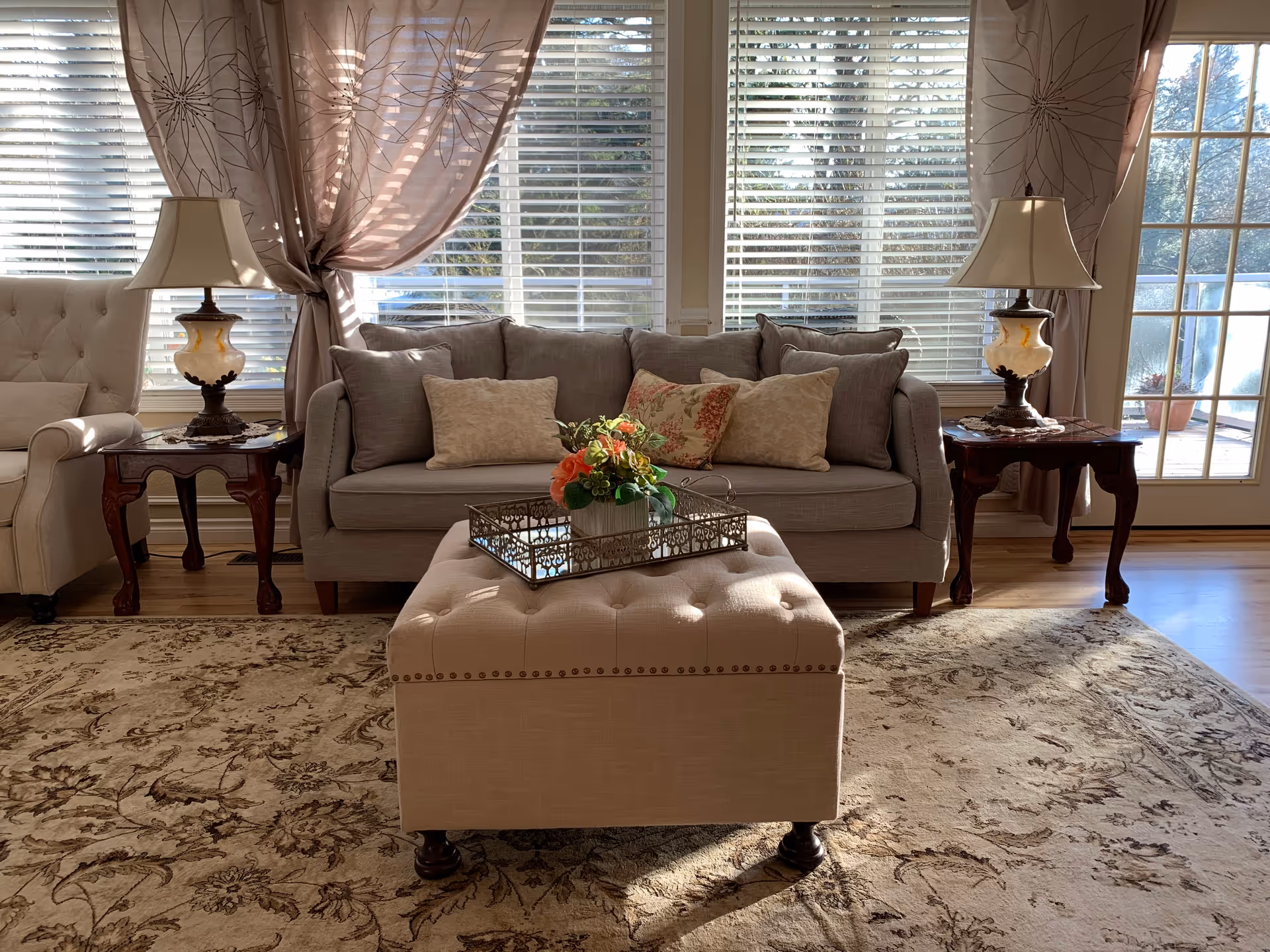 A cozy living room with a beige sofa adorned with multiple cushions, flanked by two wooden side tables each holding a decorative lamp. In front of the sofa is a tufted ottoman with a decorative tray and a flower arrangement. The room has large windows with white blinds and sheer curtains, allowing natural light to fill the space. A patterned area rug covers the wooden floor.