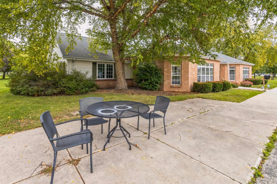 Outdoor patio with a round metal table and four chairs on a concrete walkway in front of a brick and siding single-story senior living building shaded by a large tree.