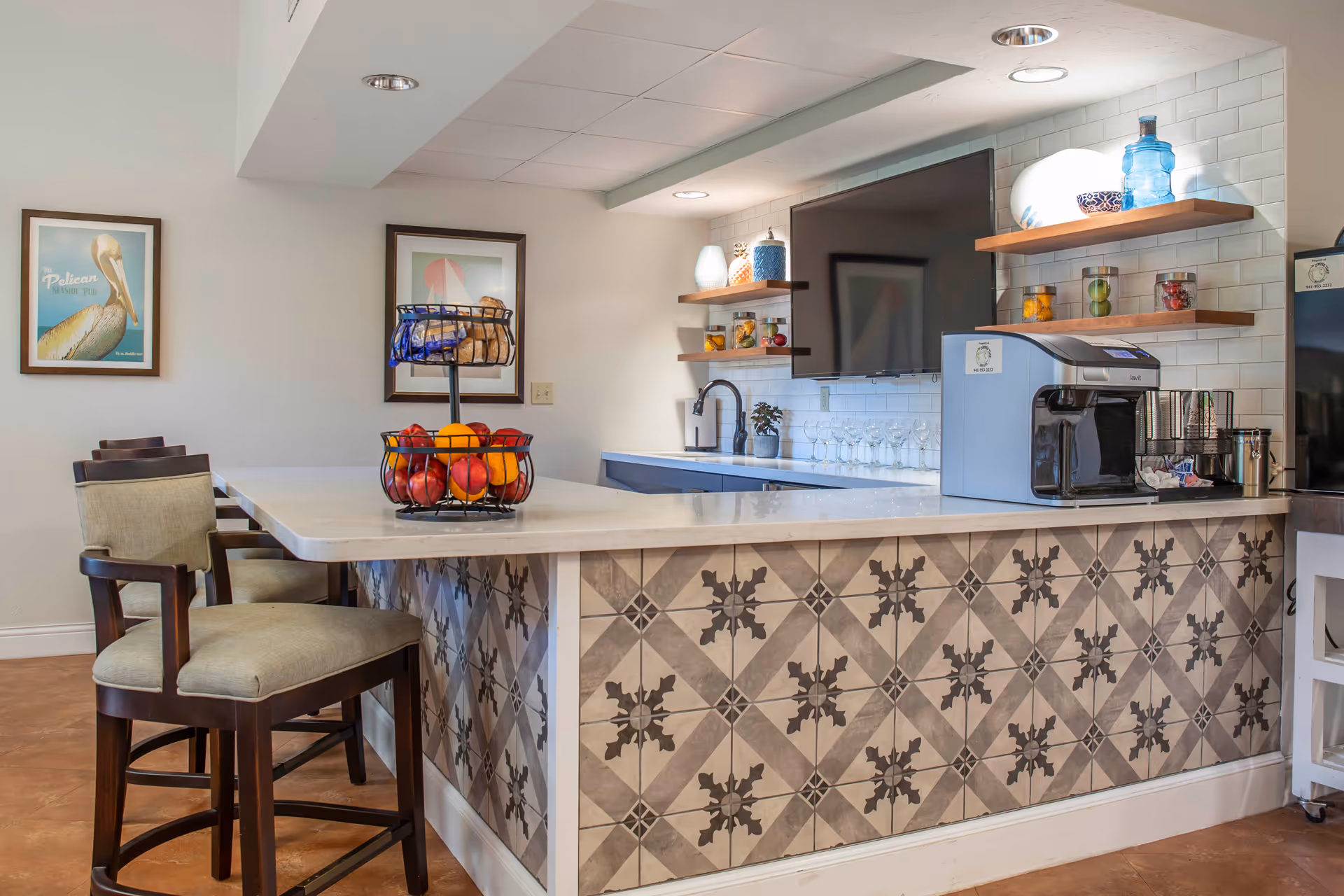 Modern kitchenette/bar area with a patterned tile counter, bar stools, fruit baskets, a coffee machine and a wall-mounted TV.