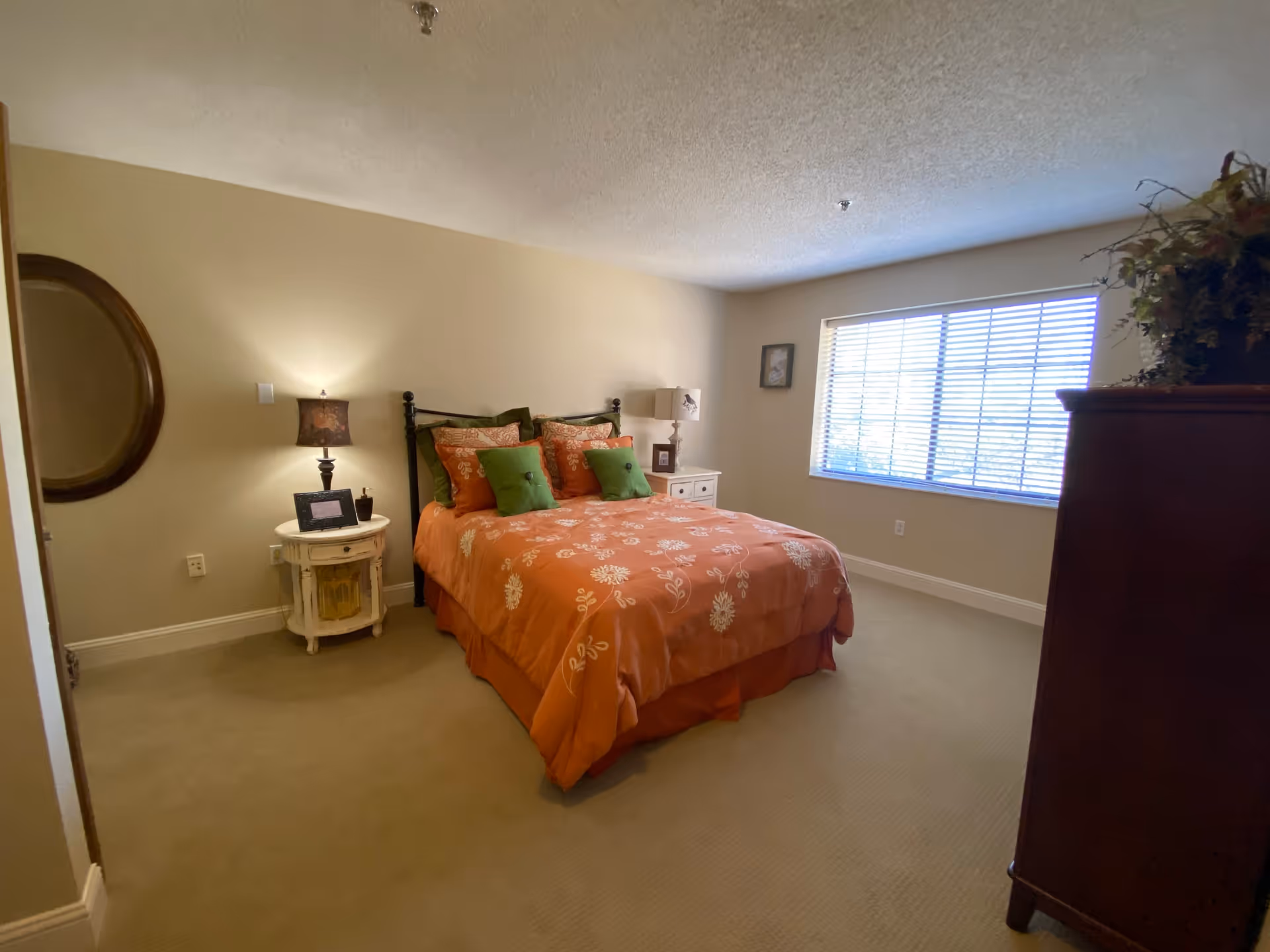 A bedroom with a bed covered in an orange floral bedspread and green pillows. There are two bedside tables with lamps on either side of the bed. A large window with blinds is on the right wall, and a round mirror hangs on the left wall. A wooden dresser with a plant on top is partially visible on the right side of the image.