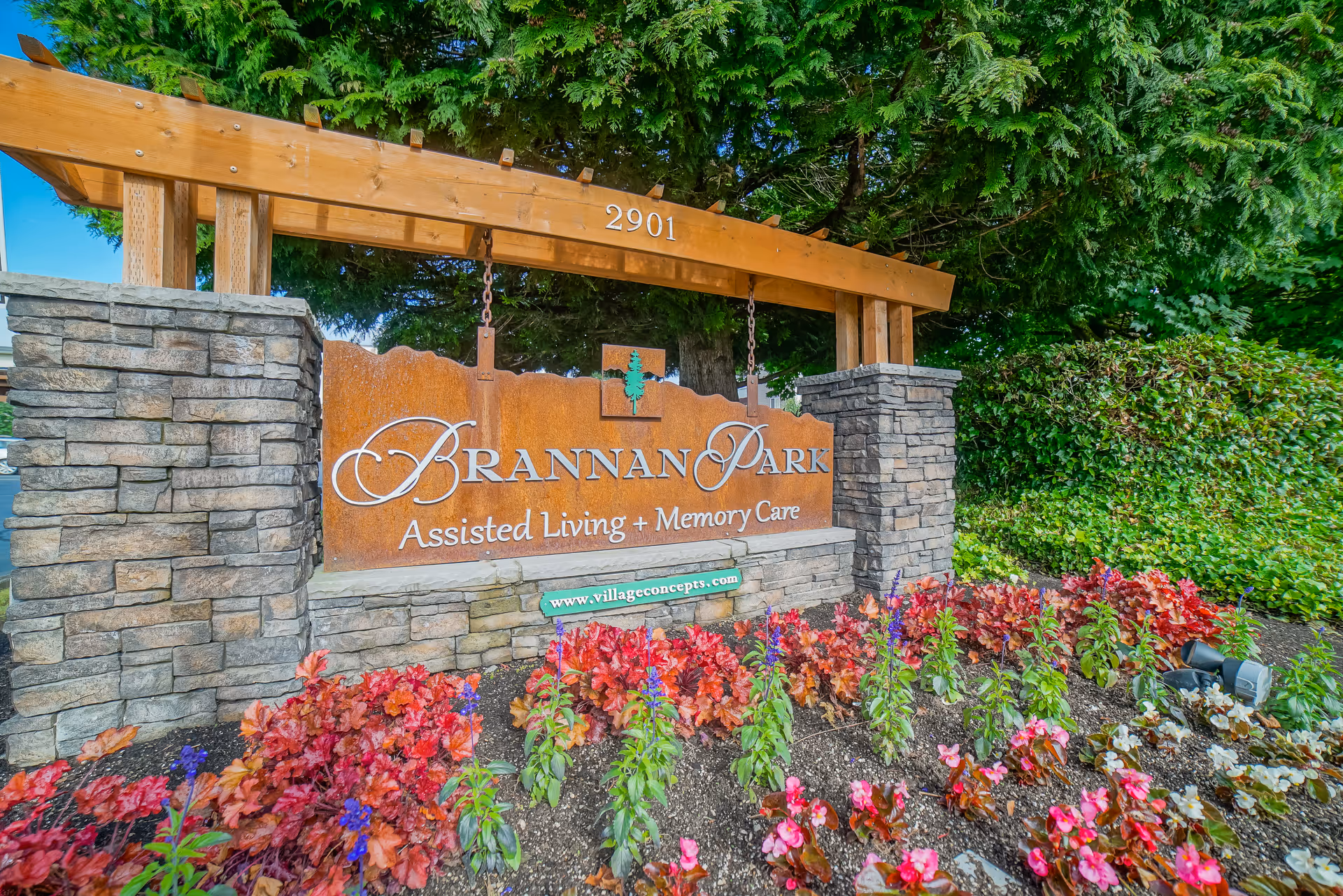 Outdoor sign for Brannan Park Assisted Living + Memory Care at 2901, surrounded by stone pillars, wooden beams, green bushes, and colorful flowers in the foreground.
