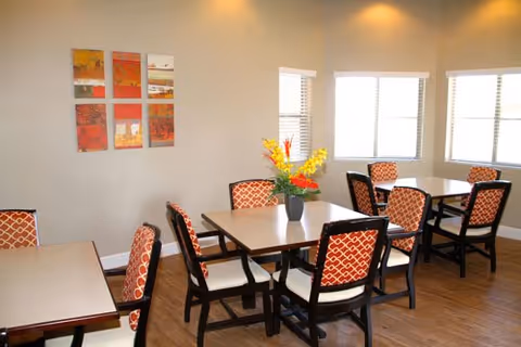 Well-lit dining room with multiple tables, patterned chairs, and a vase of flowers on a table.