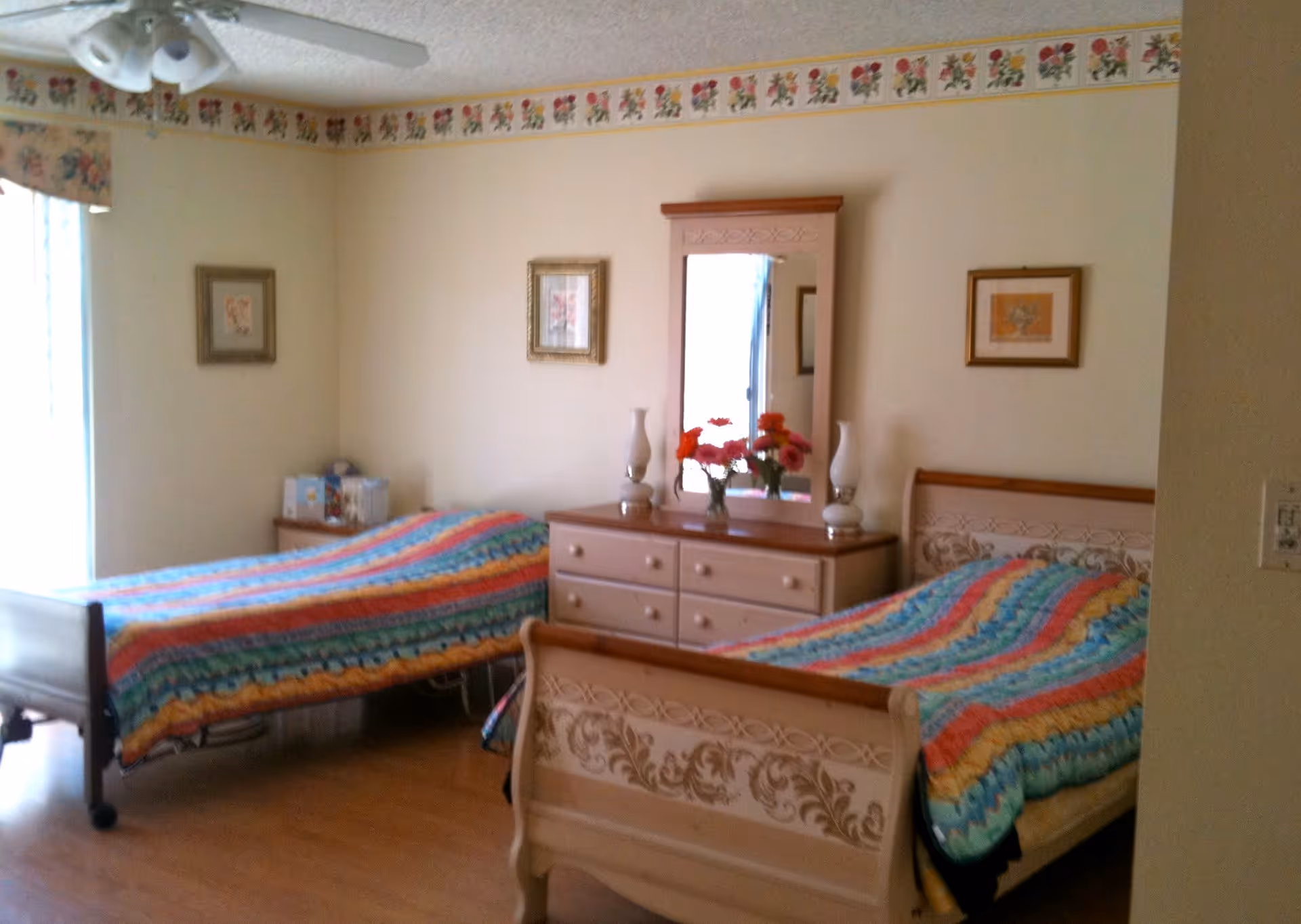 A bedroom with two single beds covered with colorful striped blankets. Between the beds is a wooden dresser with a mirror, two lamps, and a vase with flowers. The walls are light-colored with framed pictures and a decorative border near the ceiling. A ceiling fan is visible above.