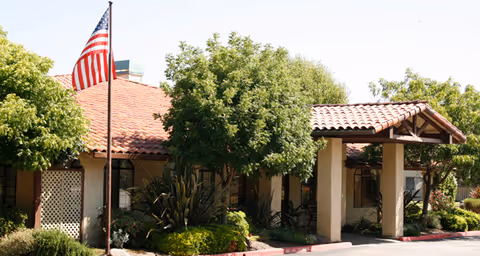 Exterior view of a single-story building with a tiled roof and a covered entrance. There are trees and shrubs around the building, and an American flag is flying on a flagpole in front.