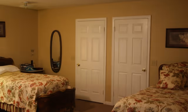 Two twin beds with floral bedspreads in a warmly lit bedroom with two closed white doors and an oval wall mirror.