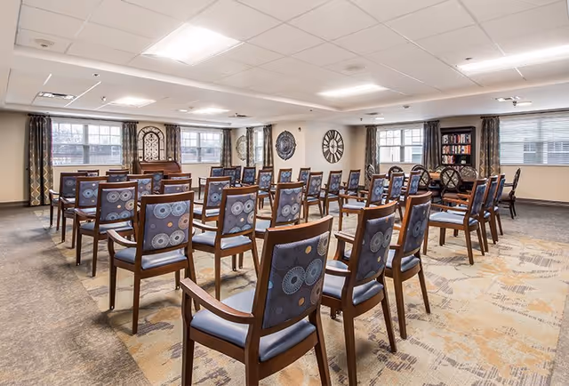 A spacious room with multiple rows of wooden chairs featuring patterned blue cushions, arranged facing a piano and a bookshelf. The room has large windows with curtains, wall clocks, and neutral-colored walls and carpet.