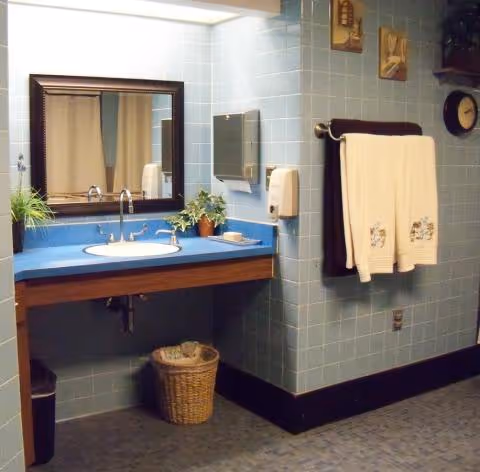 Bathroom with blue tiled walls, a sink set in a blue countertop under a mirror, towel rack with folded towels, and a wicker wastebasket.