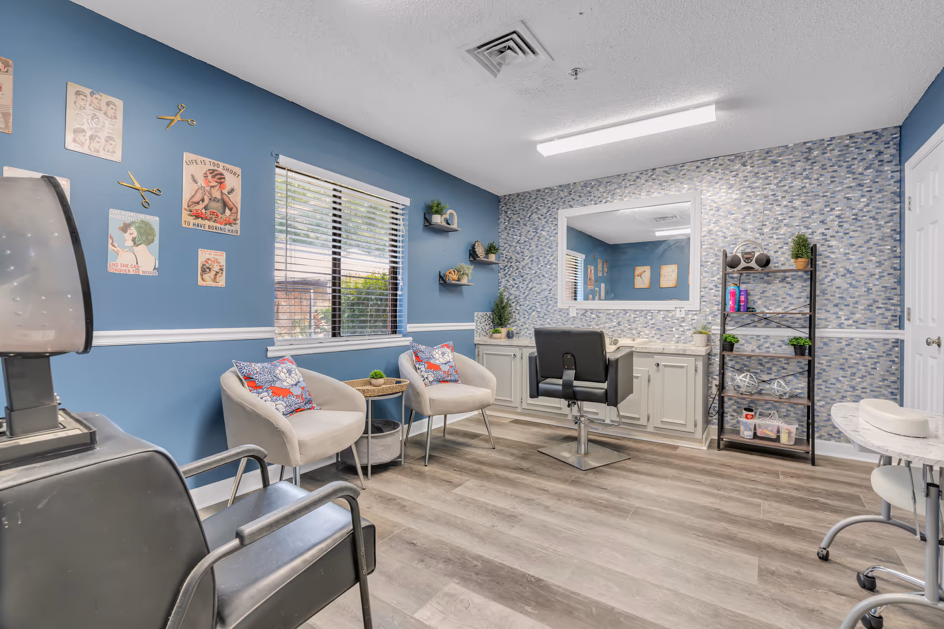 Interior of a hair salon room with blue walls and wood flooring. There are two beige chairs with colorful pillows near a window with blinds. A large mirror is mounted on a mosaic tiled wall above a white cabinet with drawers. A black salon chair is positioned in front of the mirror. A black metal shelf holds hair products and small plants. Vintage hairdressing posters and scissors decorations are on the wall.