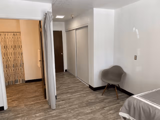 Interior view of a room with wood-patterned flooring, a gray chair against a white wall, a bed partially visible on the right, a closet with sliding doors, and an open doorway leading to a bathroom with a patterned shower curtain and a towel hanging on the wall.