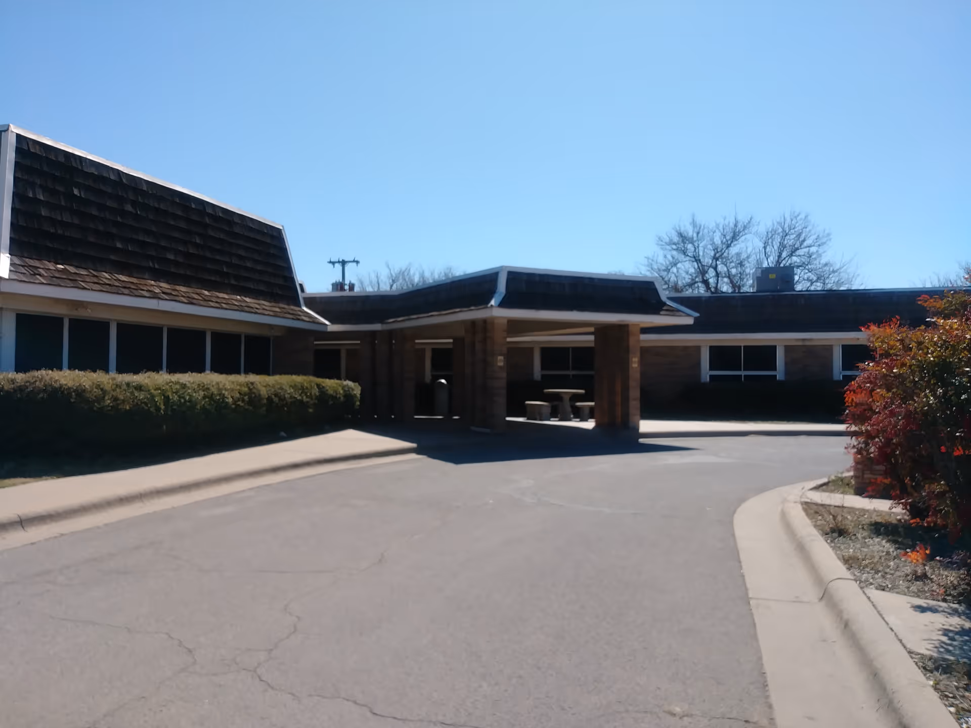 Exterior view of a single-story building with a covered entrance, surrounded by bushes and trees under a clear blue sky.