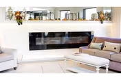 A modern living room with a white armchair on the left and a light purple sofa on the right, both with decorative pillows. A white coffee table is placed in front of the sofa. The back wall features a sleek, horizontal black electric fireplace with decorative items such as pumpkins and figurines on the mantel above it.