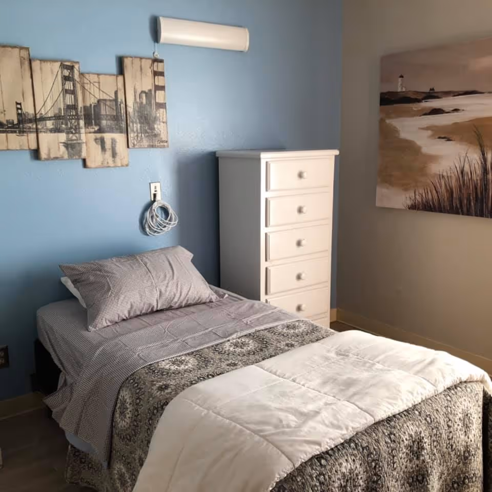 A small bedroom with a single bed made with patterned bedding and a pillow. There is a white chest of drawers next to the bed. The walls are painted blue and beige, with a multi-panel artwork of a bridge on the blue wall and a painting of a beach scene on the beige wall.
