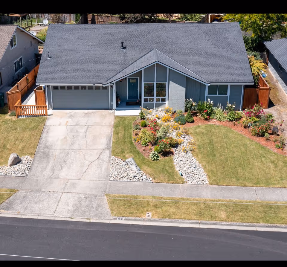A single-story house with a gray roof and blue-gray exterior. The house has a two-car garage, a front porch with a blue door, and a well-maintained front yard featuring a variety of colorful flowers and shrubs arranged around a dry creek bed made of rocks. The driveway and sidewalk are visible, along with neighboring houses and wooden fences.