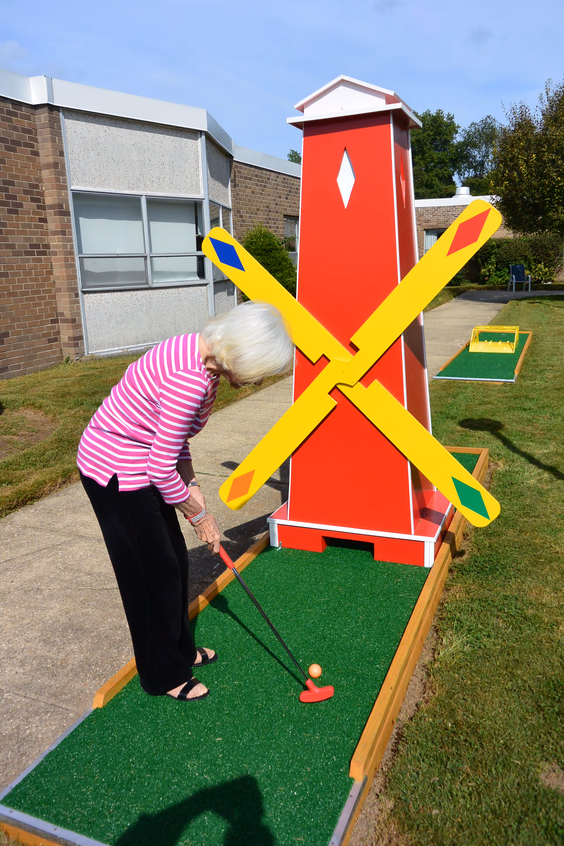An elderly woman with white hair wearing a pink and white striped long-sleeve shirt and black pants is playing mini golf outdoors. She is putting a golf ball on a green artificial turf course with a red and yellow windmill obstacle. The background shows a brick building and some greenery under a clear blue sky.