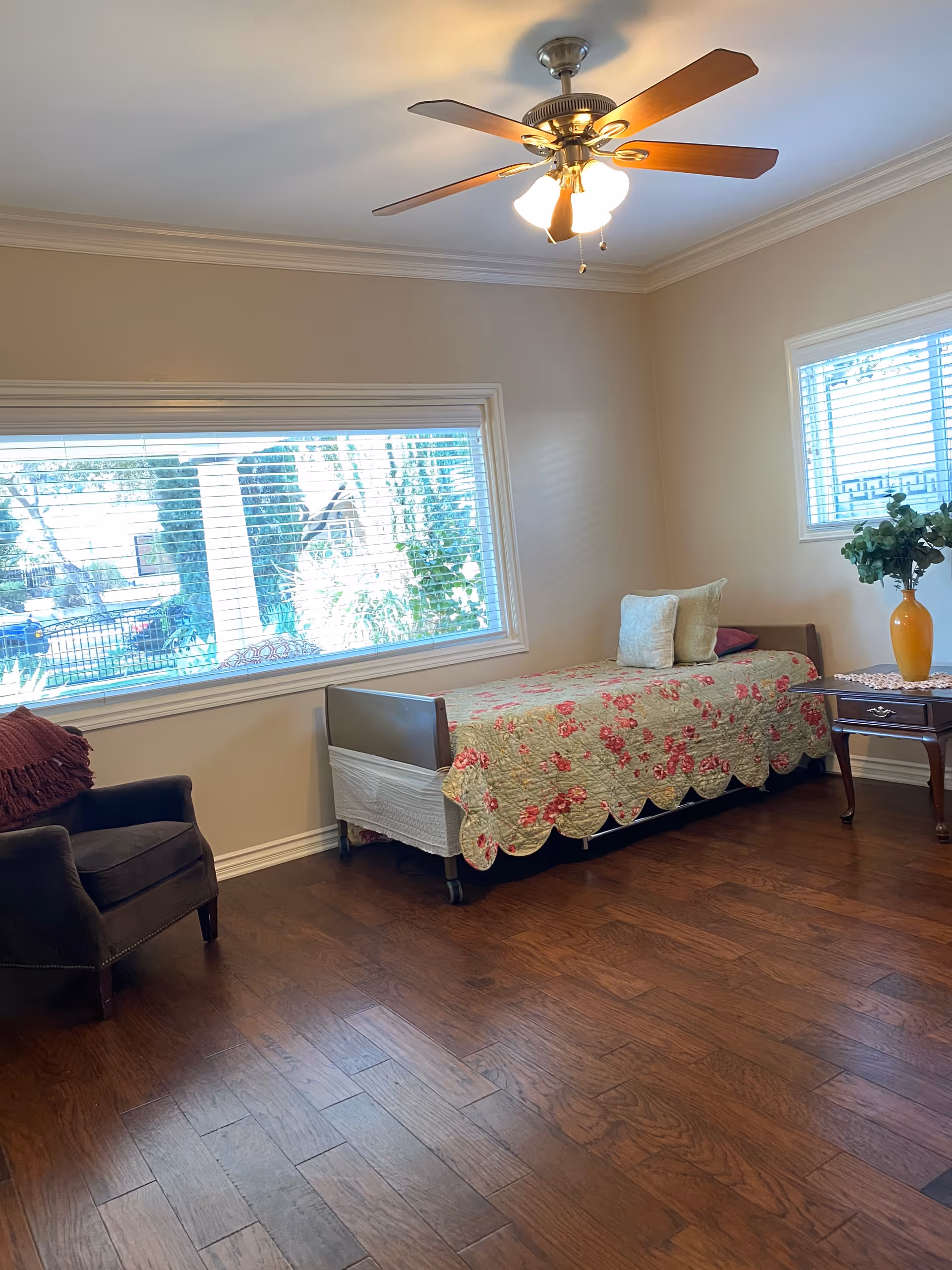 A cozy room with a single bed covered in a floral quilt, two pillows, a wooden side table with a yellow vase and green plant, a dark armchair with a red throw blanket, large windows with blinds letting in natural light, and a ceiling fan with lights.