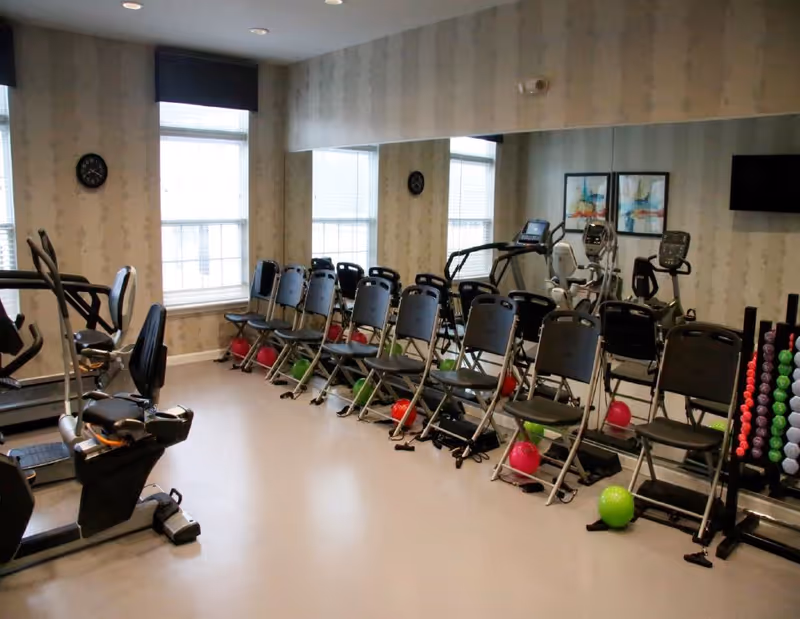Exercise room with rows of folding chairs, colorful small exercise balls, cardio machines and a rack of hand weights.