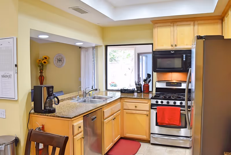 Bright L-shaped kitchen with granite counters, stainless appliances, and a red towel.