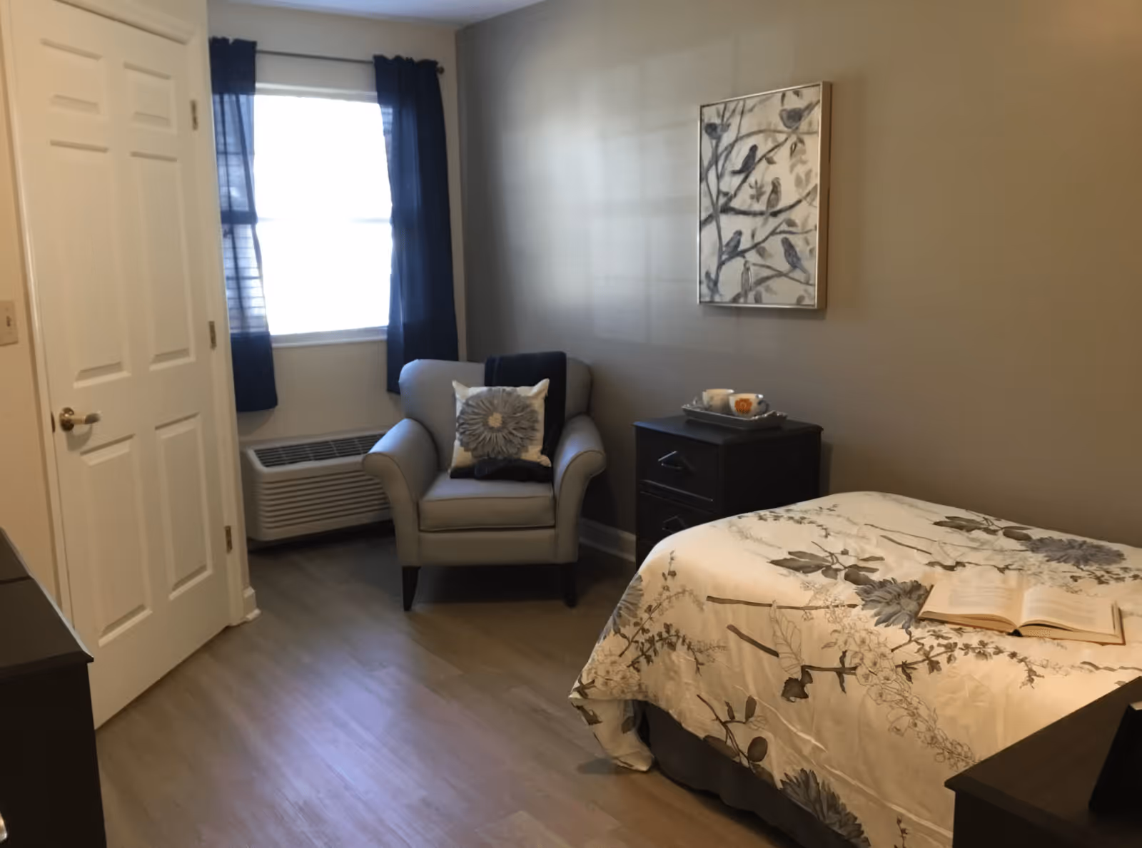 A cozy bedroom with a single bed covered in a floral-patterned comforter, an open book resting on the bed, a cushioned armchair with a decorative pillow, a small black nightstand with a tray holding a cup and a small item, a window with dark curtains, and a framed artwork of birds on branches hanging on the wall.