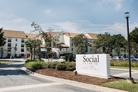 Exterior view of The Social at Savannah senior living facility with a large white sign in the foreground displaying the facility's name. The building is multi-story with a beige roof and white walls, surrounded by trees, landscaping, and a parking area under a partly cloudy sky.