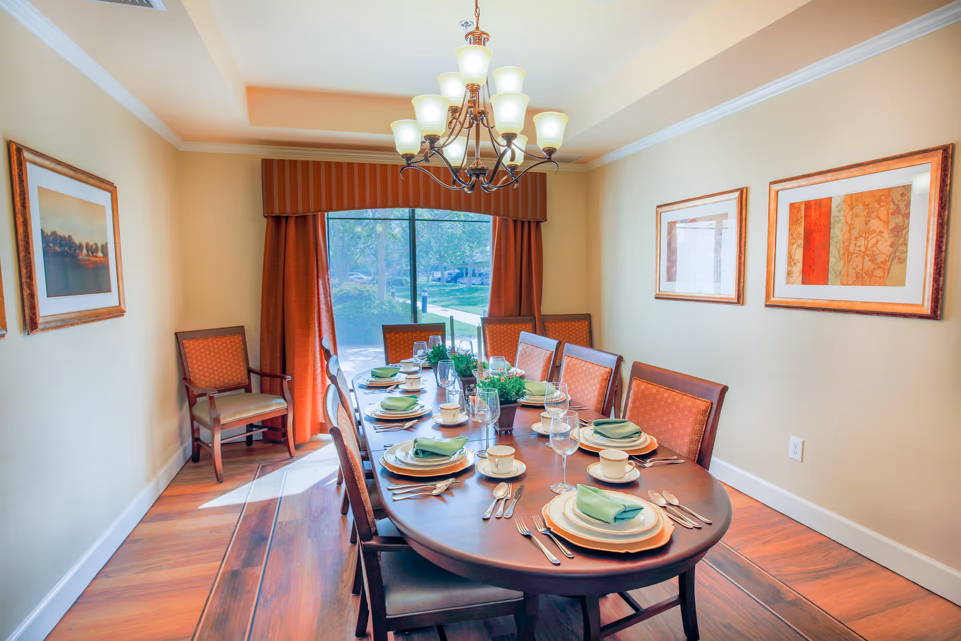A well-lit dining room with a long wooden table set for eight people. The table is arranged with plates, cups, glasses, silverware, and green napkins. There are cushioned wooden chairs around the table, and a large window with red curtains lets in natural light. The walls are decorated with framed artwork, and a chandelier with multiple lights hangs above the table.