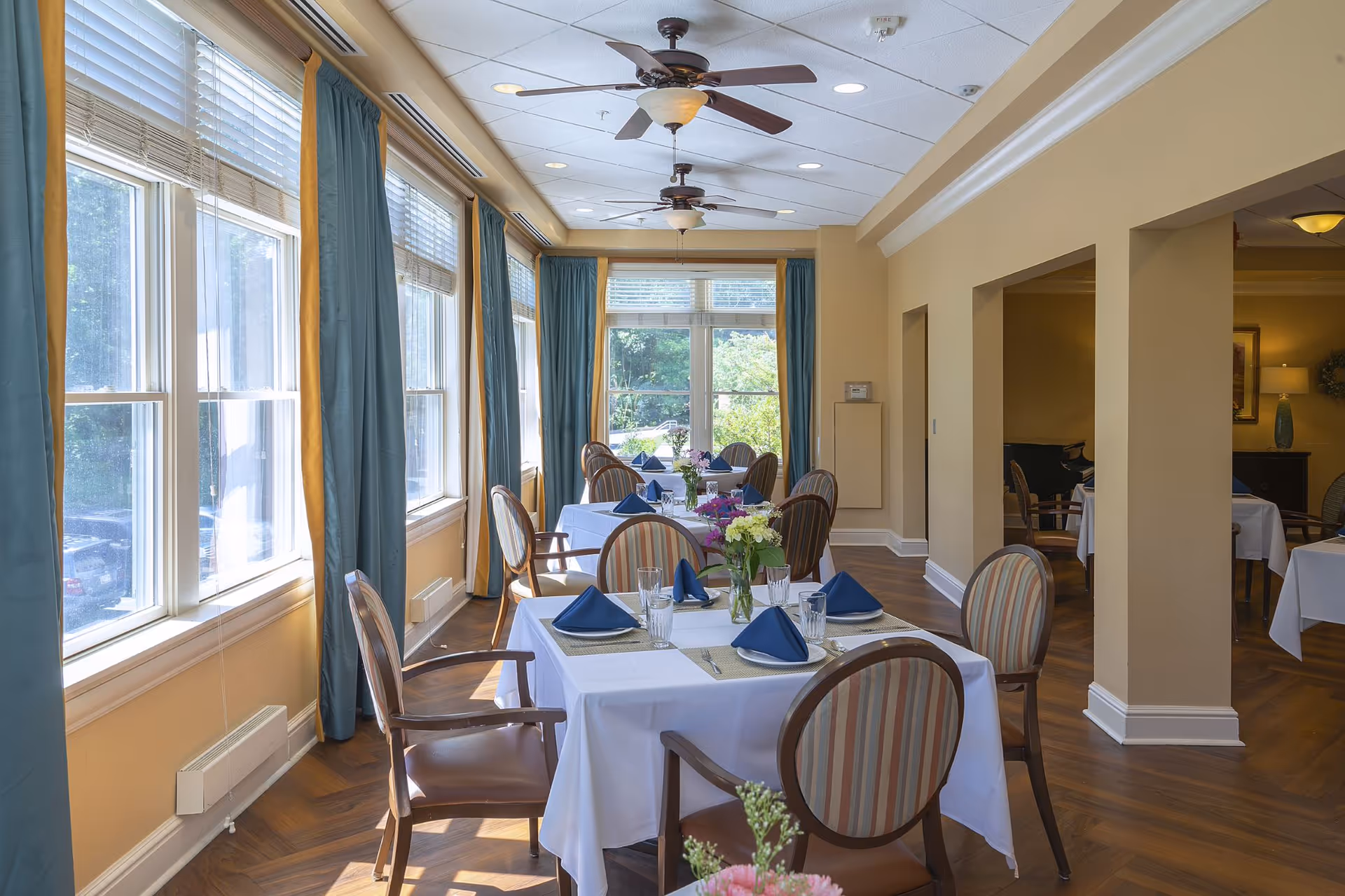 A bright dining room with large windows covered by blue and gold curtains allowing natural light to fill the space. The room features several tables covered with white tablecloths, each set with blue folded napkins, plates, glasses, and silverware. The chairs have wooden frames with striped upholstery. Ceiling fans with lights hang from the ceiling, and there are floral centerpieces on the tables.
