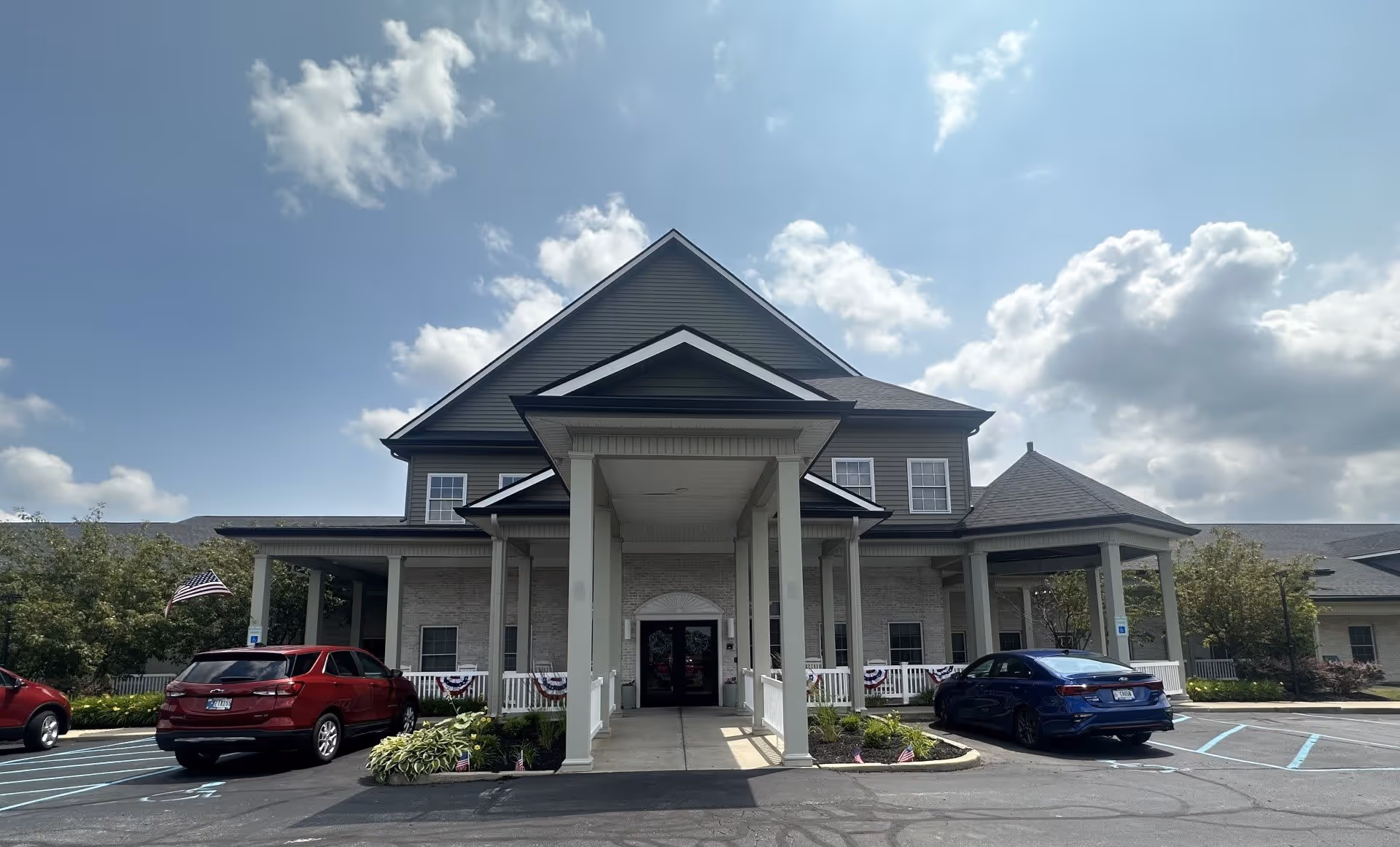 Front exterior view of a senior living facility named The Oaks at Plainfield, showing a large building with a covered entrance, several windows, and a parking area with cars. The sky is partly cloudy.