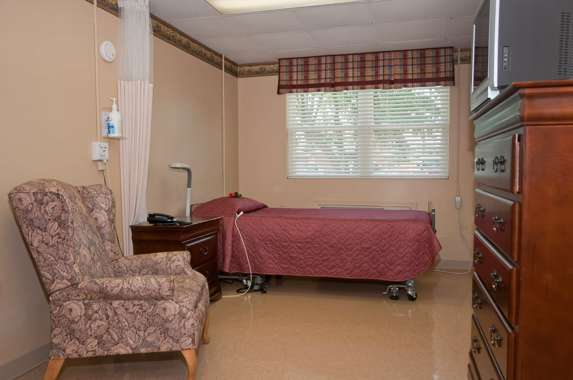 A simple bedroom in a senior living facility with a single bed covered in a maroon quilt, a floral upholstered armchair, a wooden nightstand with a lamp and phone, a large window with blinds and a plaid valance, and a wooden dresser with a television on top.