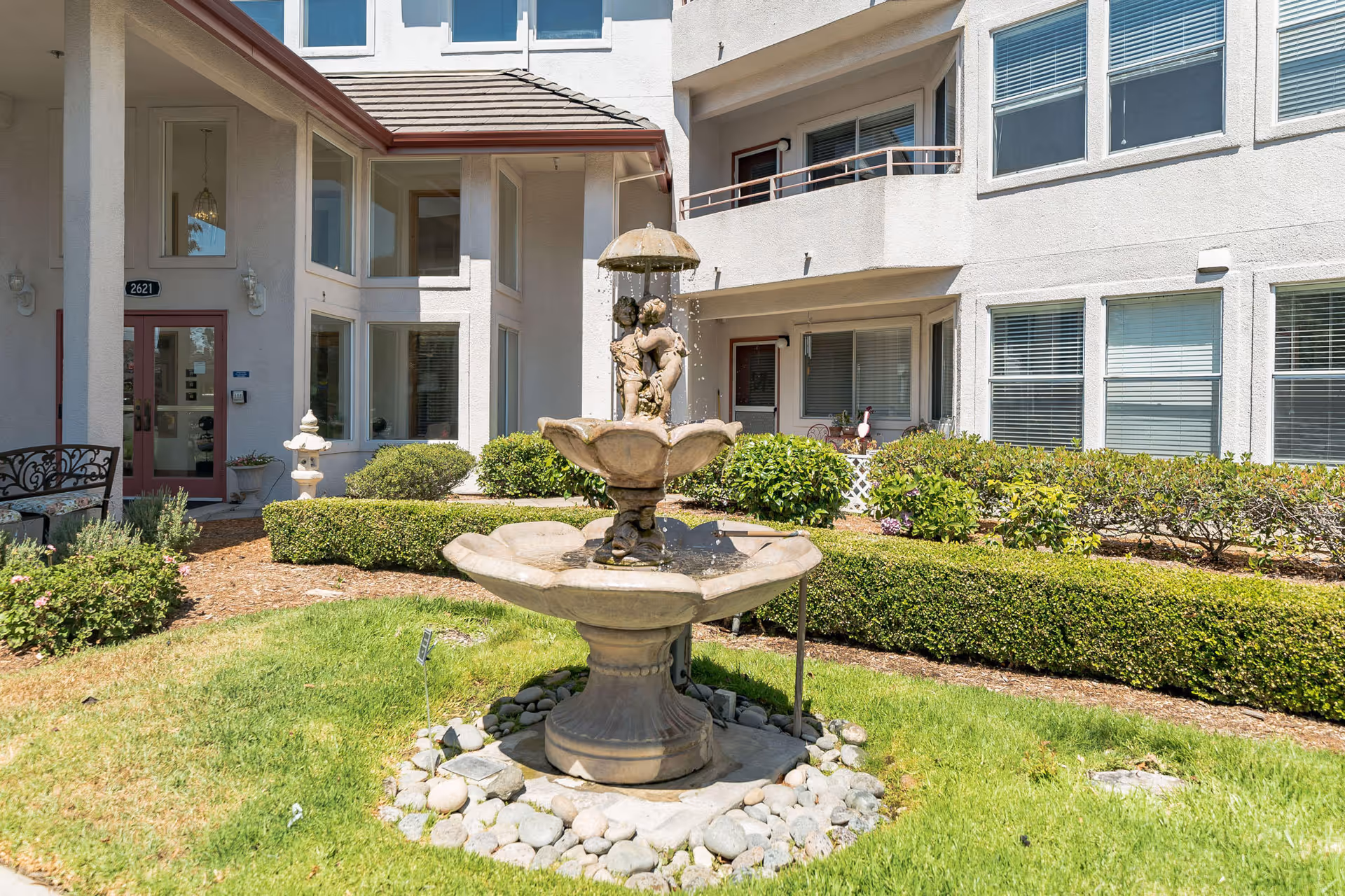 Outdoor courtyard area of Truewood by Merrill, Pinole featuring a decorative two-tier stone fountain with cherub statues in the center, surrounded by green grass, manicured bushes, and the exterior walls and windows of the building in the background.
