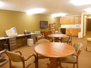 Community lounge with round tables and chairs, a kitchenette, a wall-mounted TV, and a computer desk.