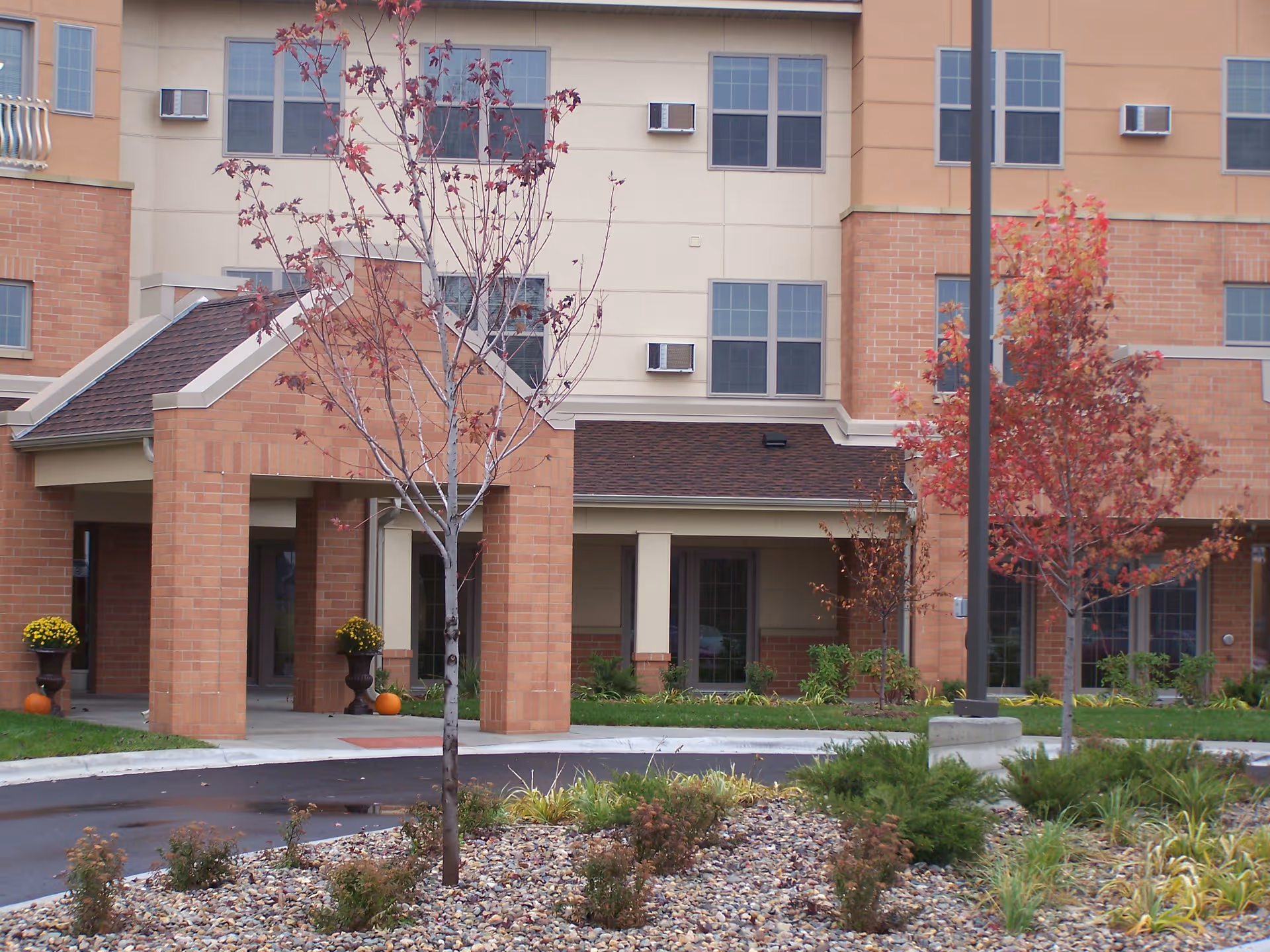 Front entrance of a brick senior living building with a covered entryway, landscaped trees, potted flowers, and pumpkins.