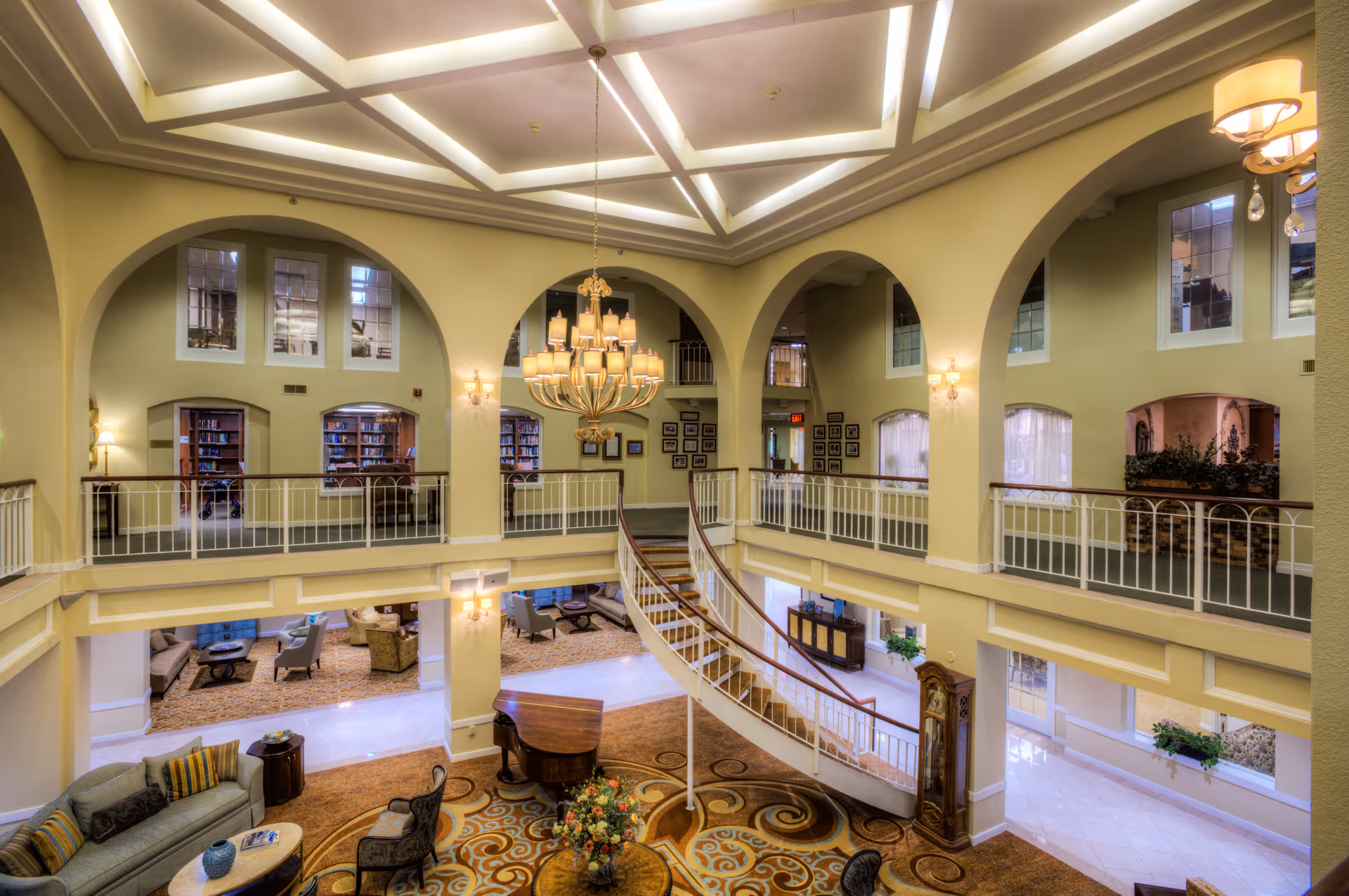 Spacious two-story living room area with a grand chandelier hanging from a coffered ceiling, a curved staircase leading to the upper level, comfortable seating arrangements including sofas and chairs, a grand piano, and large arched windows and doorways allowing natural light to fill the space.