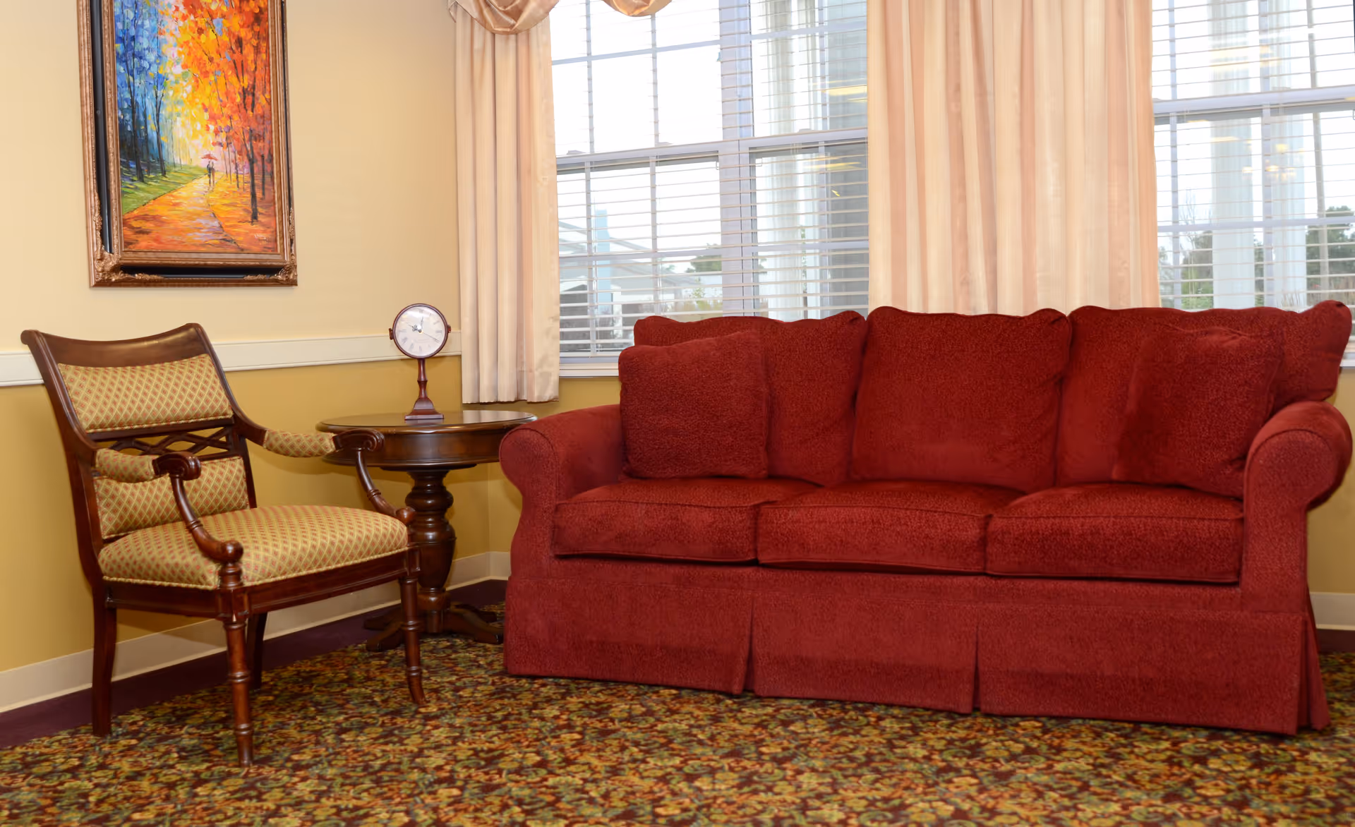 A cozy living room area with a red upholstered sofa with three cushions, a wooden side table with a small clock on it, and a wooden armchair with patterned upholstery. There is a colorful painting of a tree-lined path on the wall and large windows with beige curtains letting in natural light.