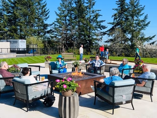 A group of elderly people sitting around a square fire pit on an outdoor patio with cushioned chairs. There are potted flowers on the patio and green grass with trees in the background. Some people are standing and walking in the background under a clear blue sky.