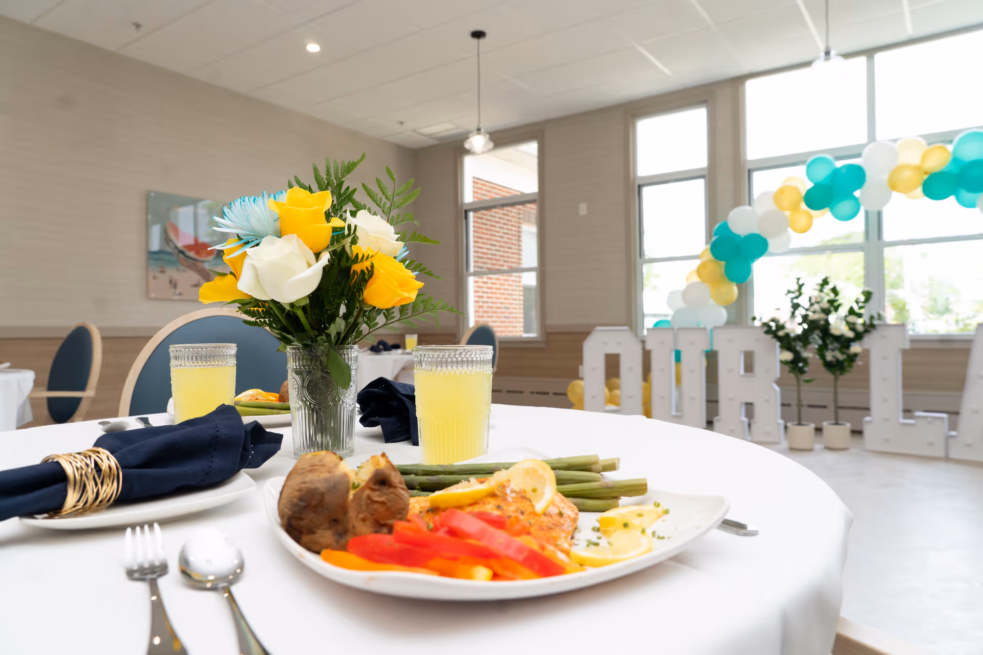 A dining table set with a plate of food including asparagus, red bell peppers, a baked potato, and a lemon garnish. Two glasses of yellow beverage and a vase with yellow, white, and blue flowers are also on the table. In the background, large windows let in natural light, and there is a balloon arch with white, teal, and yellow balloons along with large white letters spelling out 'OUR L'.