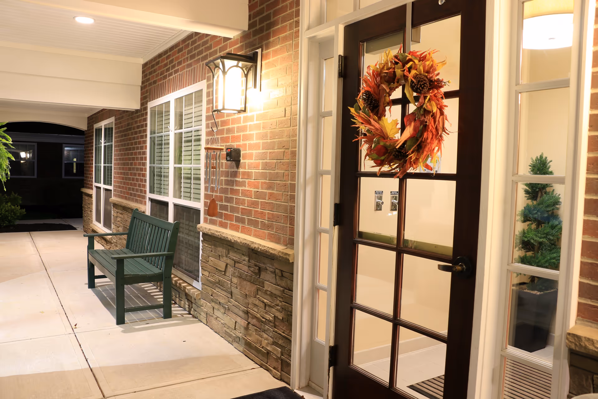 A well-lit exterior corridor of a senior living facility at night featuring a dark wooden door with glass panes decorated with an autumn-themed wreath. The brick wall has a mounted lantern light and a green bench is placed along the wall beneath windows with white frames.