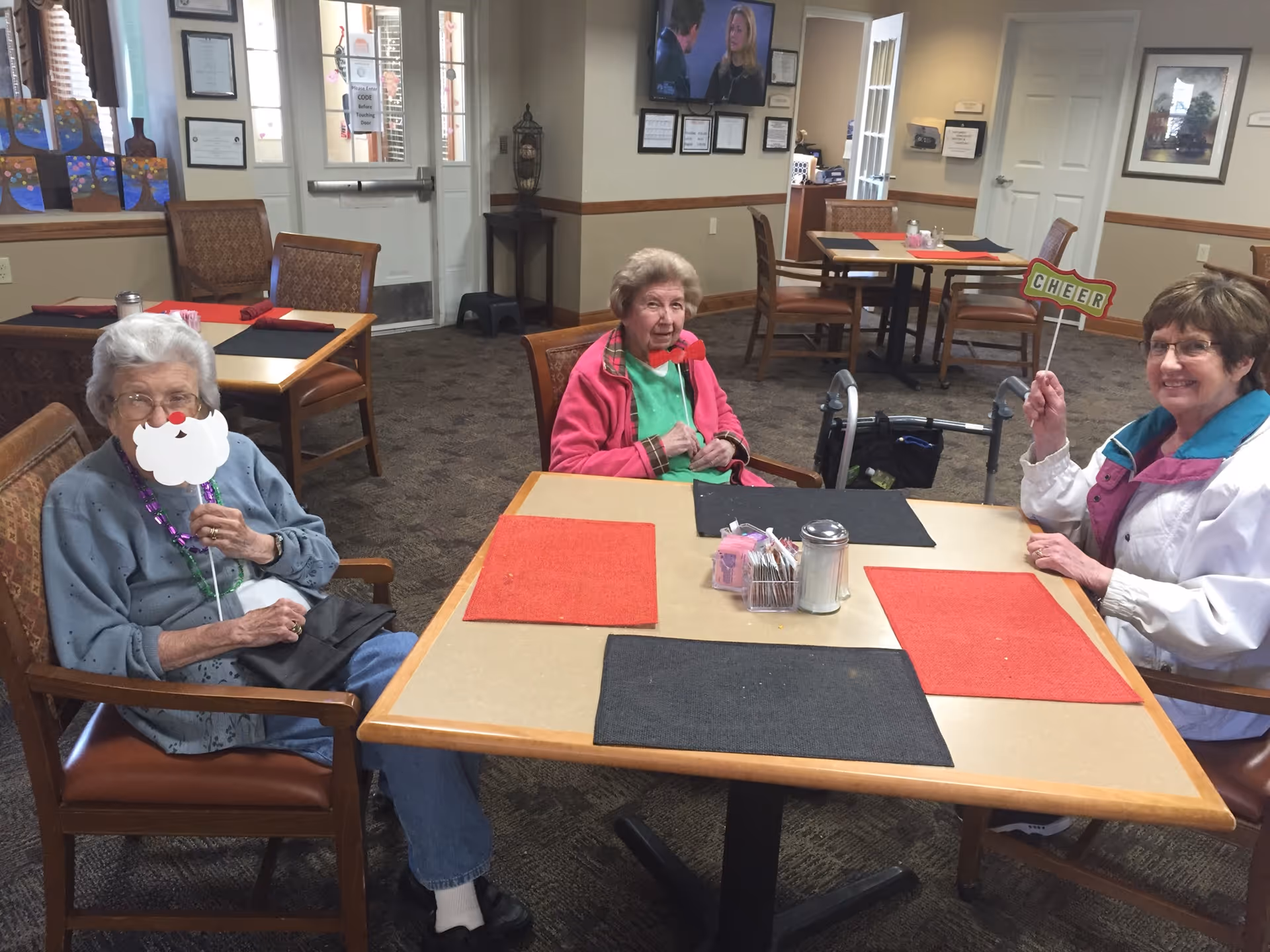 Three elderly women sit around a table in a senior living dining room, smiling and holding festive props.