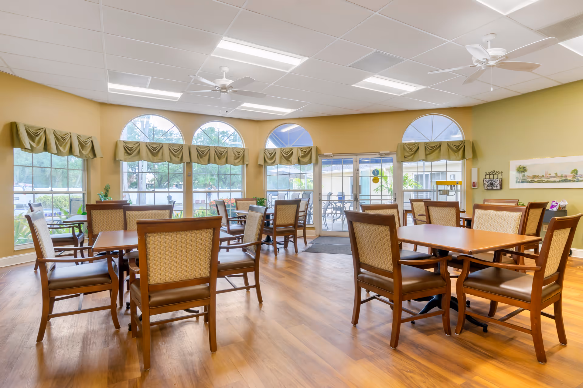 A bright dining room with wooden tables and cushioned chairs arranged neatly. Large arched windows with green valances let in natural light, and glass doors lead to an outdoor patio area. The room has a light-colored ceiling with ceiling fans and wood flooring.