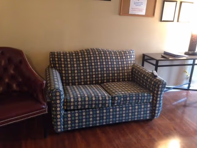 A small living area with a patterned loveseat featuring a blue and beige circular design, a dark red leather armchair to the left, and a black metal table with a lamp and some papers on the right. The floor is wooden, and the wall behind has framed certificates or documents.