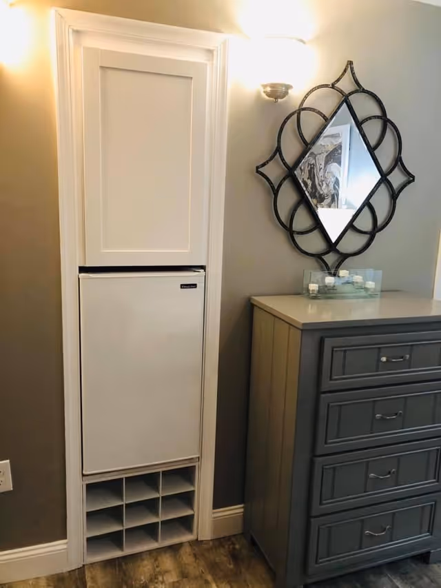 A small built-in refrigerator with a white door, set into a wall with white trim. Below the refrigerator is a cubby-style wine rack with multiple compartments. To the right is a gray chest of drawers with four drawers and silver handles. Above the chest is a decorative wall mirror with a black ornate frame and a diamond shape. The wall is painted gray, and there is a wall-mounted light fixture above the refrigerator.