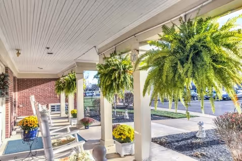 Covered outdoor porch area with white rocking chairs, hanging green ferns, and potted yellow flowers. The porch overlooks a sidewalk, parking lot, and landscaped garden beds with mulch and small shrubs.