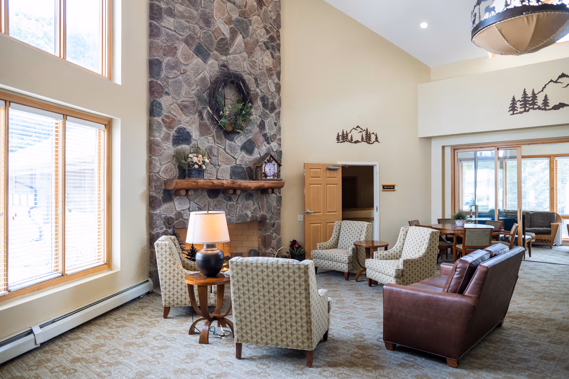 A cozy living room area with a tall stone fireplace decorated with a wreath and a clock on the mantel. The room features several patterned armchairs and a brown leather sofa arranged around a wooden side table with a lamp. Large windows with wooden frames allow natural light to fill the space. The walls are beige with rustic mountain and tree metal wall art. In the background, there is a dining table with chairs and a sliding glass door leading to another seating area.
