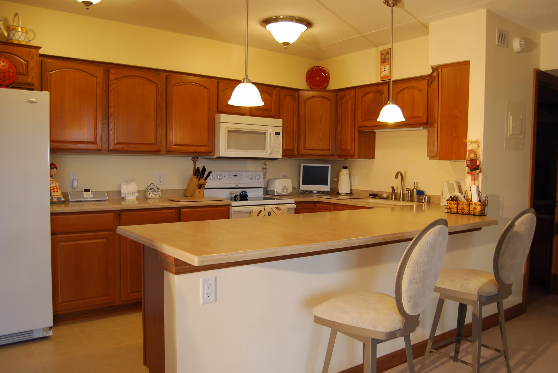 A kitchen with wooden cabinets, a white refrigerator, a white stove with an overhead microwave, and a countertop with two cushioned bar stools. The kitchen is well-lit with two hanging pendant lights and a ceiling light fixture. Various kitchen items and decorations are placed on the countertops and cabinets.