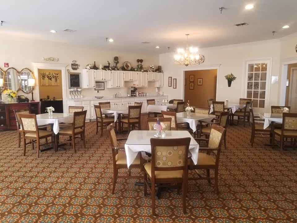 A dining room with multiple square tables covered with white tablecloths, each surrounded by four wooden chairs with patterned cushions. The room features a patterned carpet, white walls, a chandelier, and a kitchenette area with white cabinets and decorative items on top. There are also mirrors, framed pictures, and a small American flag on one of the tables.