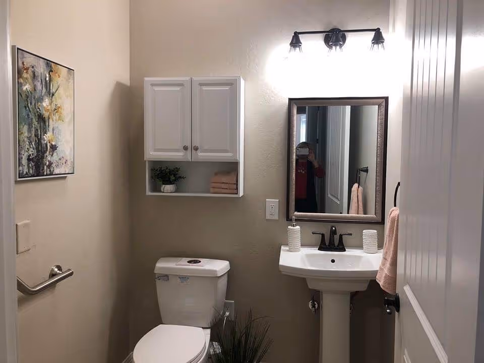 A small bathroom with a white toilet and a white pedestal sink. Above the sink is a rectangular mirror with a dark frame and a three-light fixture. To the left of the toilet is a white wall cabinet with two doors and an open shelf holding a small plant and folded towels. A floral painting hangs on the left wall, and a pink towel is hanging on a ring next to the sink. The walls are painted beige.