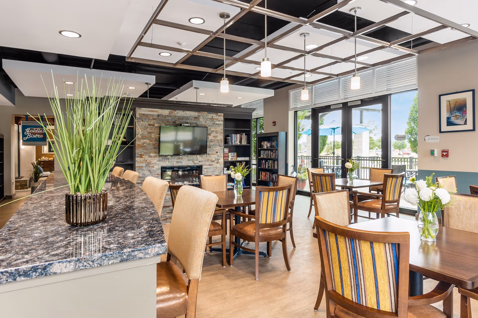 A bright and modern dining area with wooden tables and chairs, some with striped upholstery. A granite countertop with a decorative plant is in the foreground. The room features a stone accent wall with a mounted flat-screen TV and a fireplace below it. Large windows and glass doors provide a view of an outdoor patio with umbrellas. Shelves with books and board games are visible near the TV. Pendant lights hang from the ceiling, and fresh flowers in vases decorate the tables.